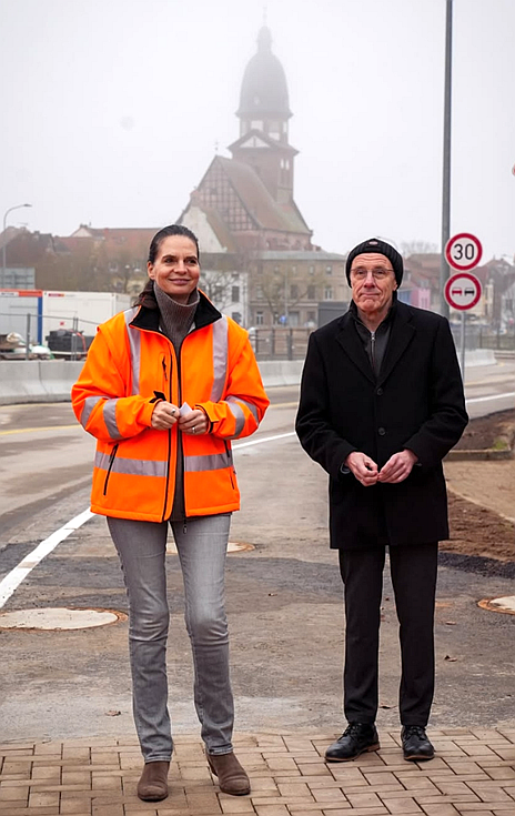 Ines Jesse und Norbert Möller stehen vor dem fertigen Teilstück der neuen Brücke. Im Hintergrund sind die Baustelle und die Kirche von Waren (Müritz) in Nebel eingehüllt.