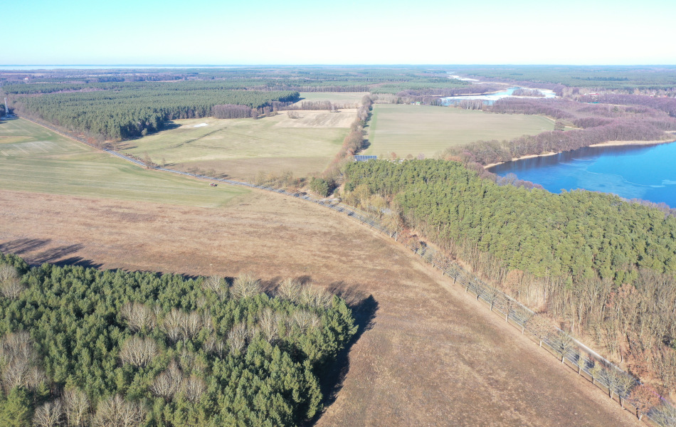 Blick von oben über Wälder und Felder. Eine Straße führt quert durch die Felder und an einem Wald vorbei, an dem sich ein See befindet.