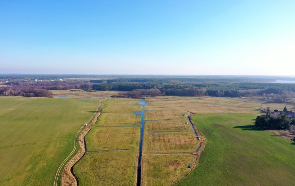 Blick von oben über Felder und Wälder. Mehrere kleine Bäche/Flüsse führen über ein Feld. Weiter rechts davon befinden sich Häuser.