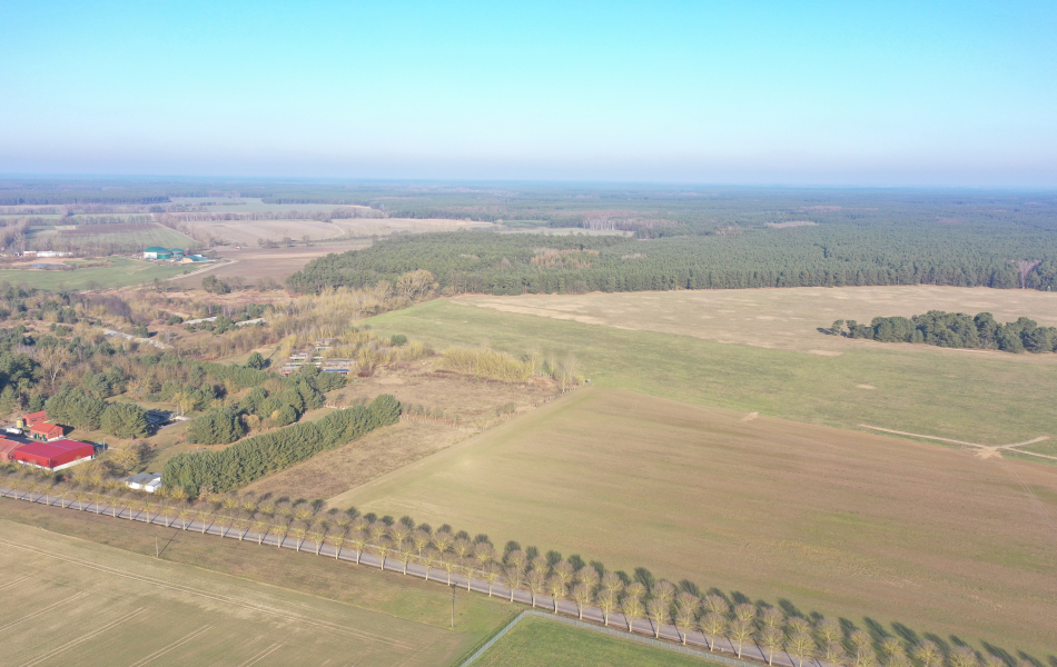 Blick von oben über Felder und Wälder. Auf der linken Seite befinden sich mehrere Gebäude an den Feldern. 