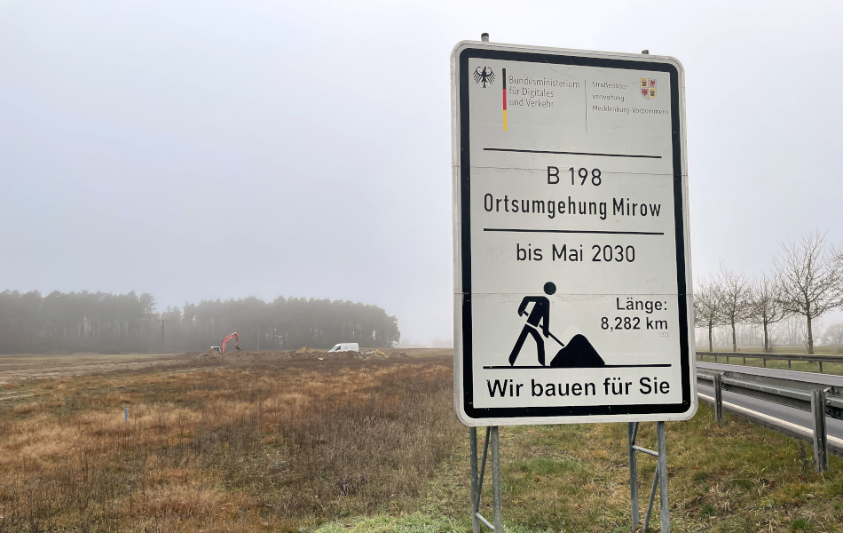 Baustellenschild zur B 198 Ortsumgehung Mirow am Straßenrand; dahinter Nebel, Felder und Waldrand.