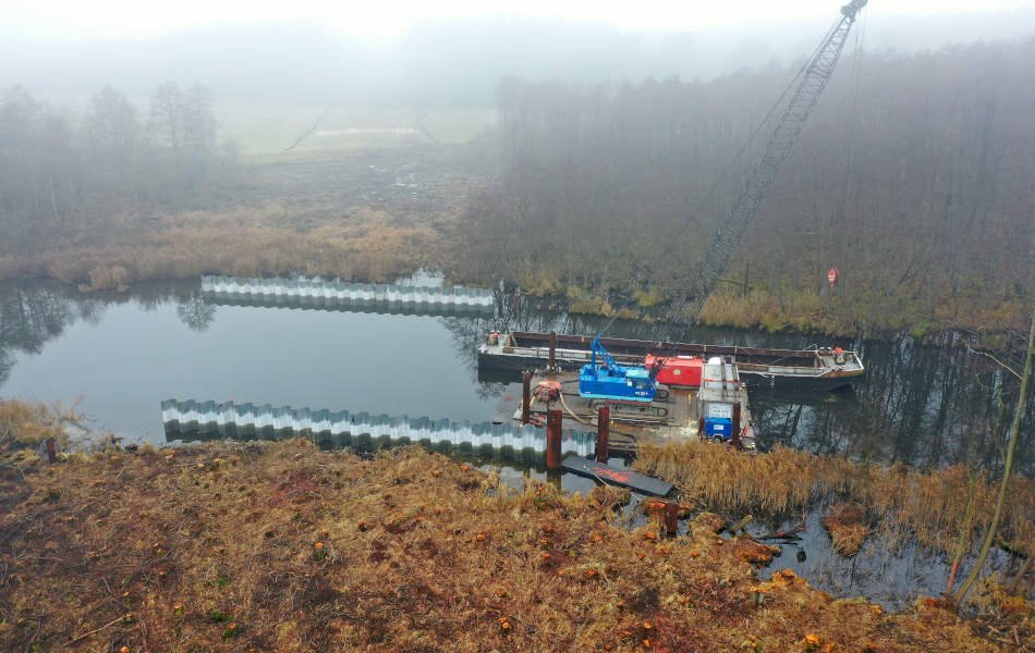 Baustelle einer Brücke über ein Gewässer mit Ponton, Kran und Spundwänden in offener Niederungslandschaft.