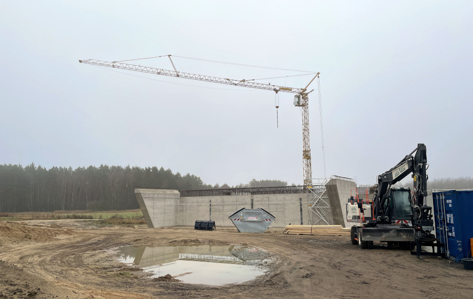 Baustelle mit Brückenbauwerk aus Beton und großem Turmkran; rechts Bagger und Container, davor nasser Baugrund mit Pfützen, im Hintergrund Wald unter nebligem Himmel.