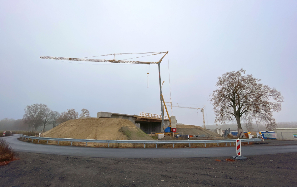Baustelle der Ortsumgehung Mirow an der Lärzer Straße mit Brückenbauwerk, Erddamm und Baukran; im Vordergrund kurvige Fahrbahn mit Schutzplanken, im Hintergrund Nebel über Feldern und Baumreihen.