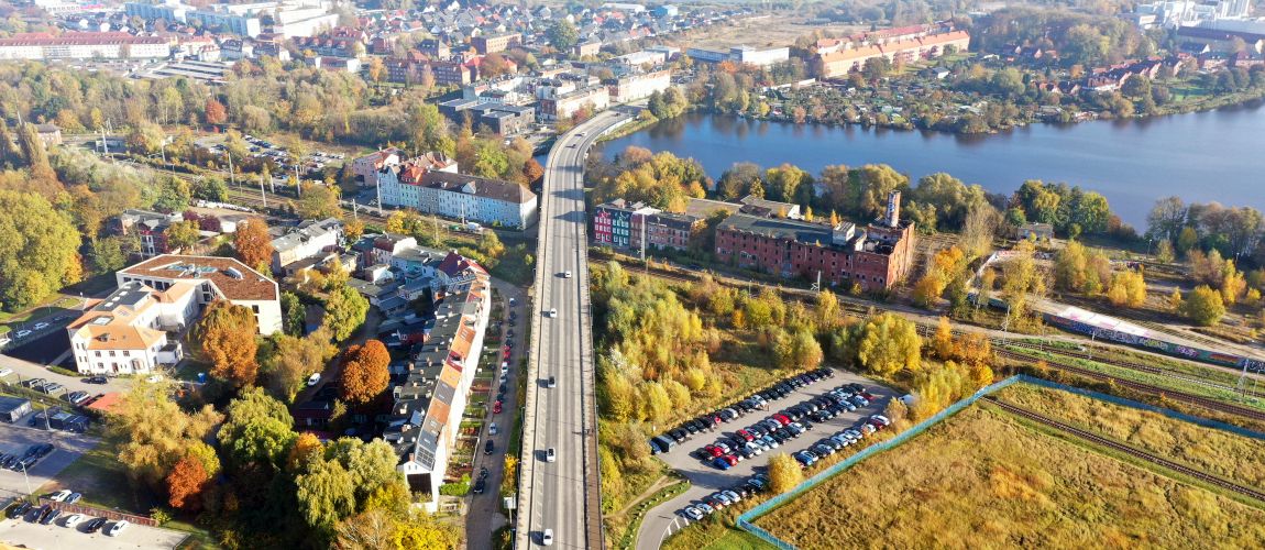 Mehre Autos fahren über eine Straßenbrücke, die durch ein Stadtgebiet und über ein Gewässer führt. Neben der Brücke sind Wohnhäuser und Parkplätze.