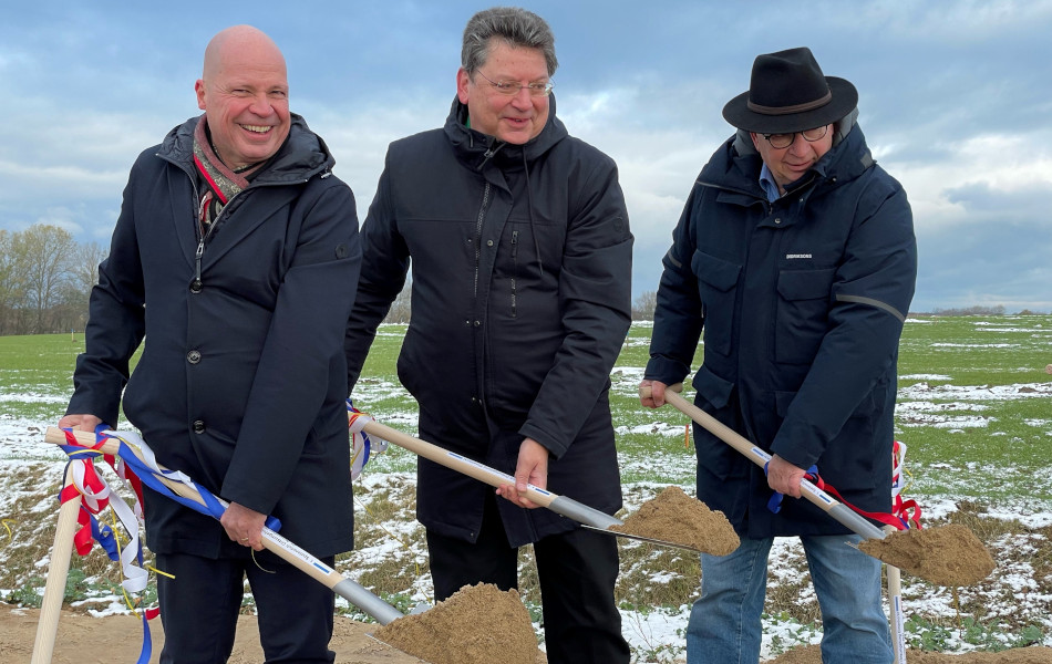 Drei Männer stehen nebeneinander, lächeln und heben jeweils mit beiden Händen einen Spaten voll Erde in die Luft. Im Hintergrund ist ein Feld, vereinzelt bedeckt mit Schnee.