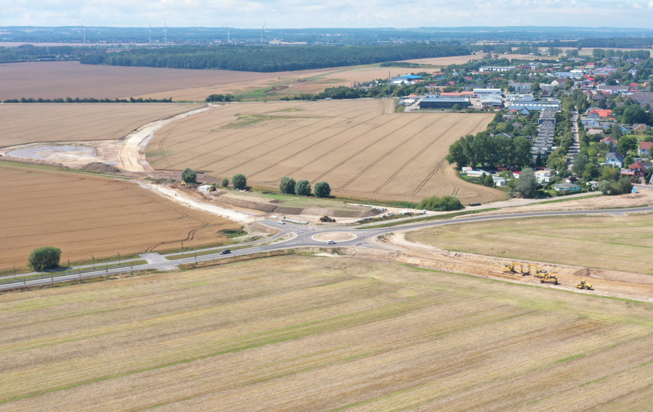 Blick von oben auf einen Kreisverkehr, der insgesamt vier Zufahrten hat. Zwei davon sind mit Baustellenbaken abgesperrt, weil die Straßen hier gerade gebaut werden. Sandtrassen auf Felder deuten den künftigen Verlauf der Straßen an. Eine der beiden bereits fertiggestellten Ausfahrten führt zum Ort Elmenhorst.