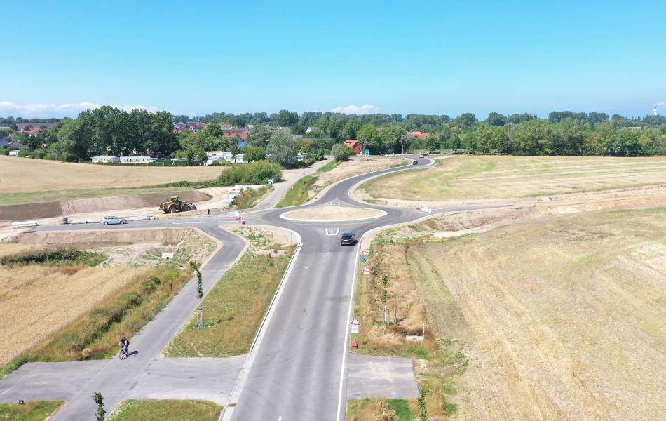 Blick von oben auf eine Straße, die auf einen Kreisverkehr zuläuft. Der Kreisel hat vier Ausfahrten, aber es geht nur geradeaus weiter, weil die Ausfahrten nach links und rechts sind mit Baustellenbaken abgesperrt. Die Straßen hier werden gerade noch gebaut.