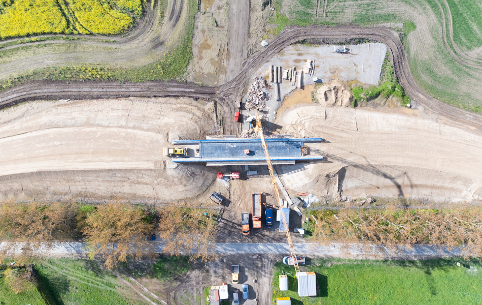 Blick von oben auf eine Brücke, die sich im Bau befindet. Unter der Brücke befinden sich viele Baufahrzeuge. Auf der Brücke fährt ein Radlader. Rampen aus Erde führen links und rechts auf die Brücke.