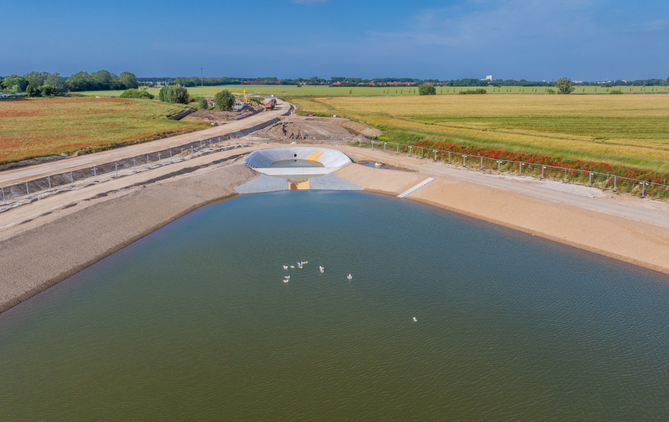 An einer Sandtrasse auf einem Feld liegt ein großes Becken, das mit Wasser gefüllt ist. Auf dem Wasser schwimmen Schwäne.