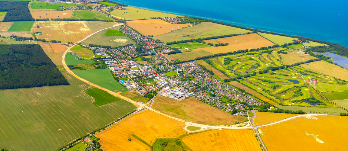 Auf einer Luftaufnahme ist der Ort Elmenhorst, eingebettet in einer Felderlandschaft an der Ostsee, abgebildet. In einem Halbkreis liegt die neue Ortsumgehungstrasse, überwiegend aus Sandboden bestehen, um den Ort herum.