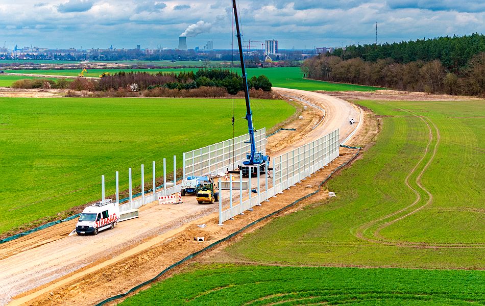 Eine Sandtrasse führt durch Felder hindurch. Im Hintergrund sind Wälder. Auf der Trasse sind Baufahrzeuge aktiv. Es werden große Wandelemente an den Seiten der Erdtrasse eingehoben. © Pixelbox15 Eine Sandtrasse führt durch Felder hindurch. Im Hintergrund sind Wälder. Auf der Trasse sind Baufahrzeuge aktiv. Es werden große Wandelemente an den Seiten der Erdtrasse eingehoben.