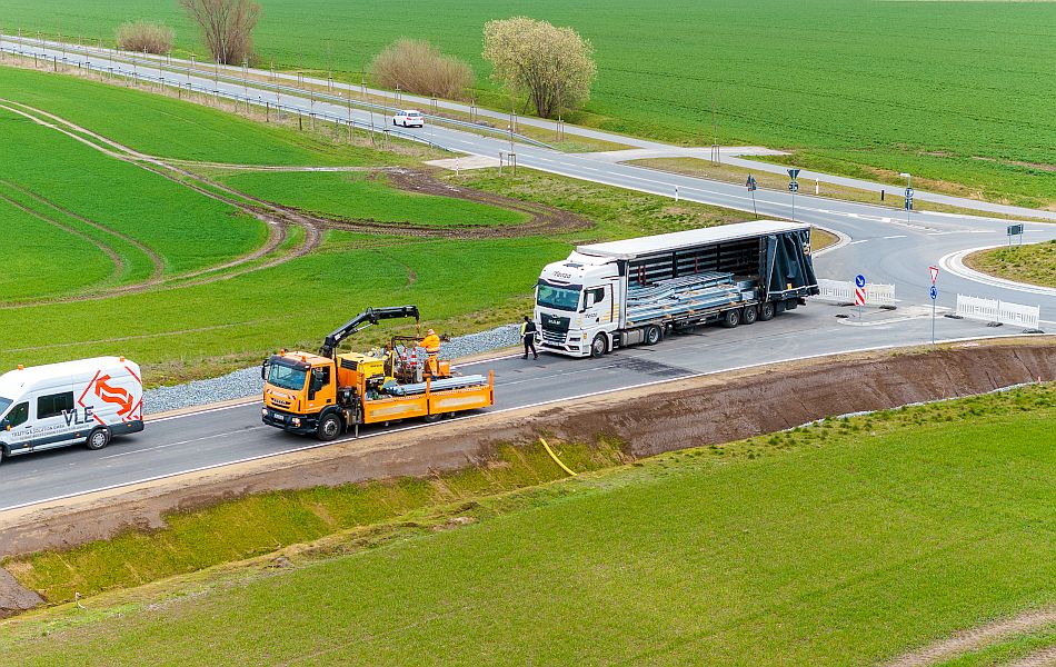 Ein Lkw liefert Material für den Aufbau von Schutzplanken an. Davor steht ein Baufahrzeug mit einem Kran, um das Material an der neuen Straße zu verteilen. © Pixelbox15 Ein Lkw liefert Material für den Aufbau von Schutzplanken an. Davor steht ein Baufahrzeug mit einem Kran, um das Material an der neuen Straße zu verteilen.