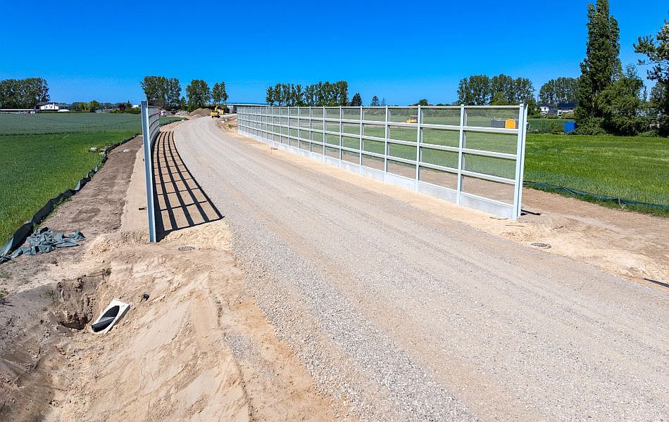Eine Trasse aus Erde und Schotter führt durch zwei Felder hindurch. Im Hintergrund ist ein Bagger auf der Trasse. An der Trasse befinden sich auf jeder Seite zwei große, lange Wände, die aus Glaselementen bestehen. © Pixelbox15 Eine Trasse aus Erde und Schotter führt durch zwei Felder hindurch. Im Hintergrund ist ein Bagger auf der Trasse. An der Trasse befinden sich auf jeder Seite zwei große, lange Wände, die aus Glaselementen bestehen.