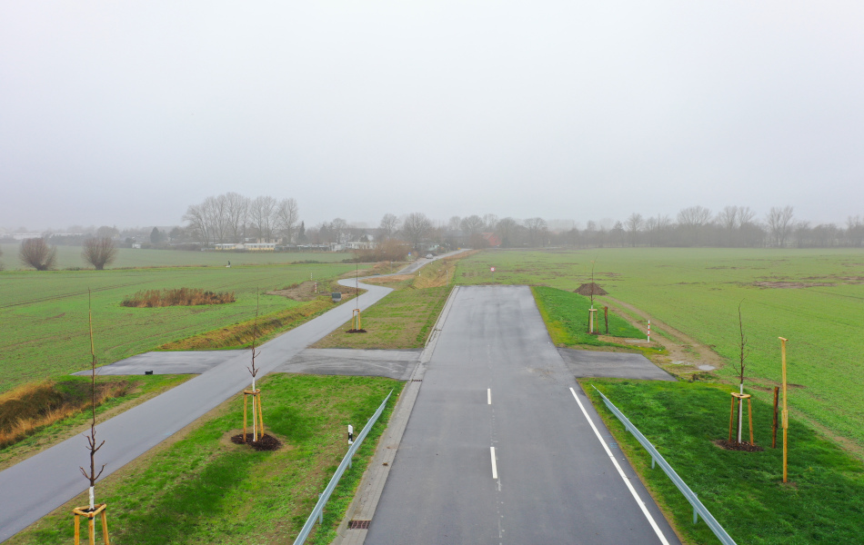 Blick von oben auf eine neu gebaute Straße, die von Schutzplanken und frisch gepflanzten Bäumen gesäumt wird. Die Straße endet auf einem Feld. Links neben der Straße führt ein asphaltierter Geh- und Radweg über das gesamte Feld entlang.