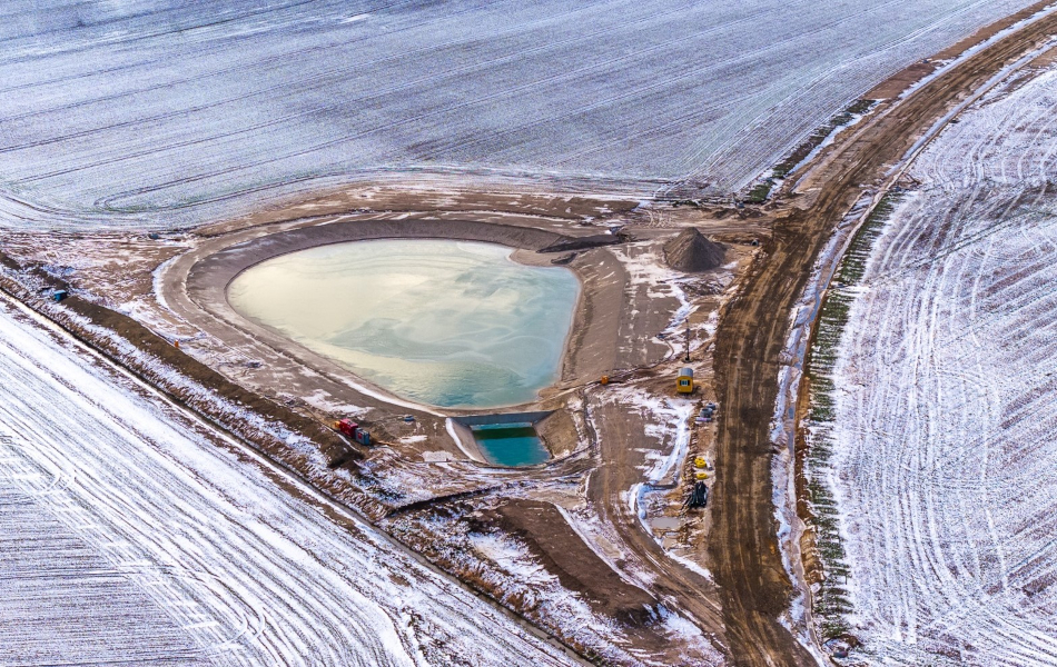 An einer Trasse auf einem Feld liegt ein großes Becken, das mit Wasser gefüllt ist. Auf dem Feld liegt Schnee.