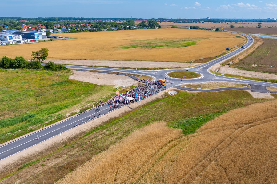 Viele Menschen stehen auf einer neuen Straße. Sie feiern die Eröffnung. Die Straße führt zu einem neuen Kreisverkehr auf dem Land.