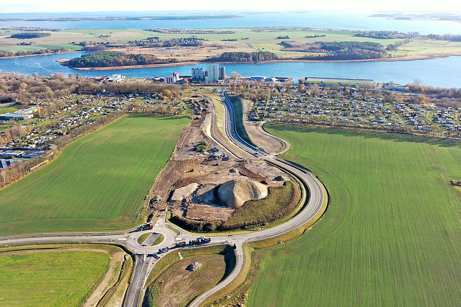 Luftbild eines Kreisverkehrs mit angeschlossenem Neubauabschnitt der Straße, dahinter grüne Felder, Baustellenflächen und das Peenestrom-Ufer.