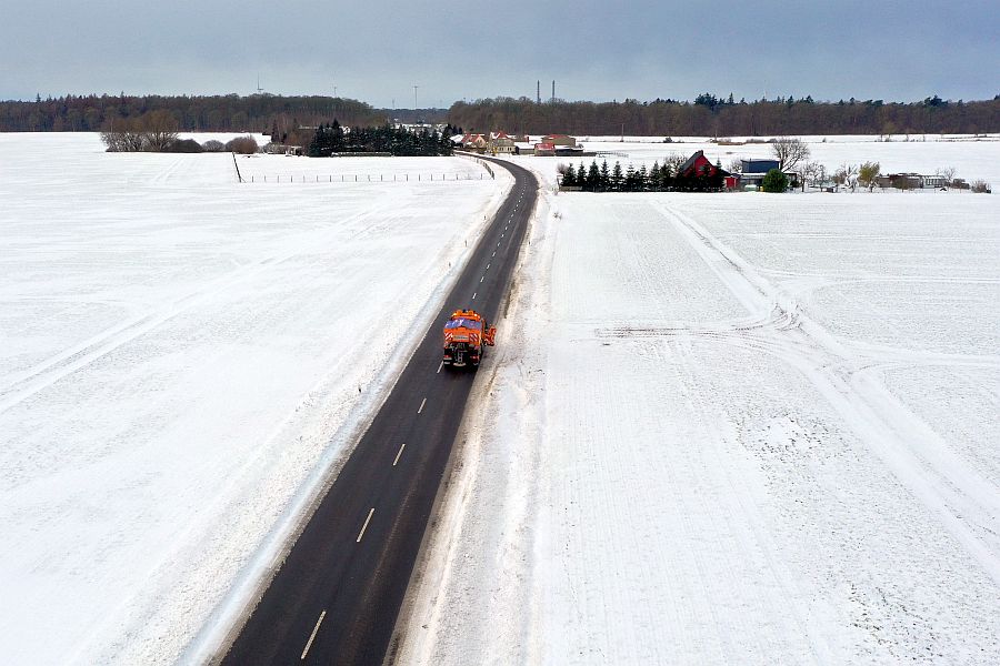 Luftaufnahme einer geräumten Landstraße durch verschneite Felder; ein orangefarbener Winterdienst-Lkw fährt Richtung Ortschaft, im Hintergrund Wald und grauer Himmel.