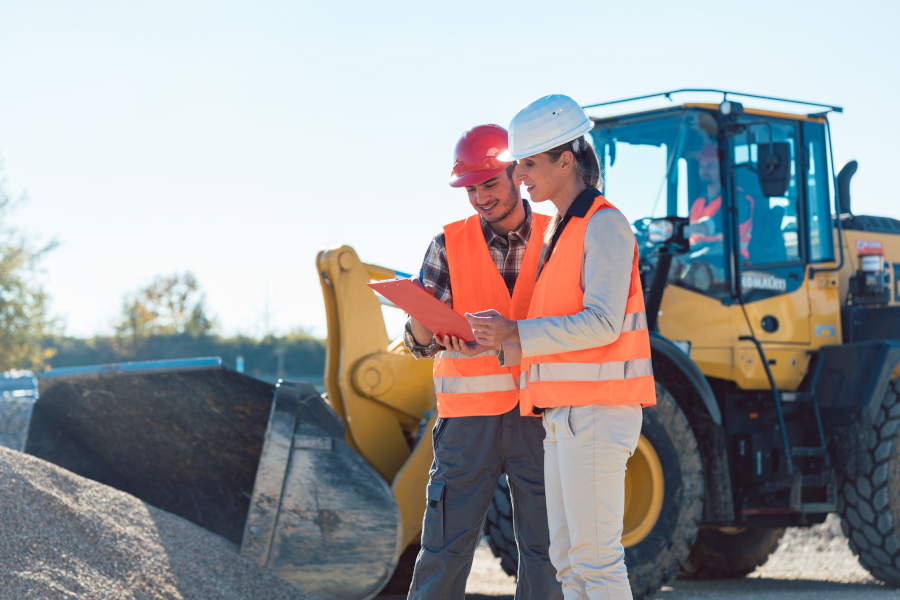 Ein Mann und eine Frau stehen auf einer Baustelle und schauen auf einen Plan. Sie tragen orangene Westen und Bauhelme. Im Hintergrund ist ein Radlader.