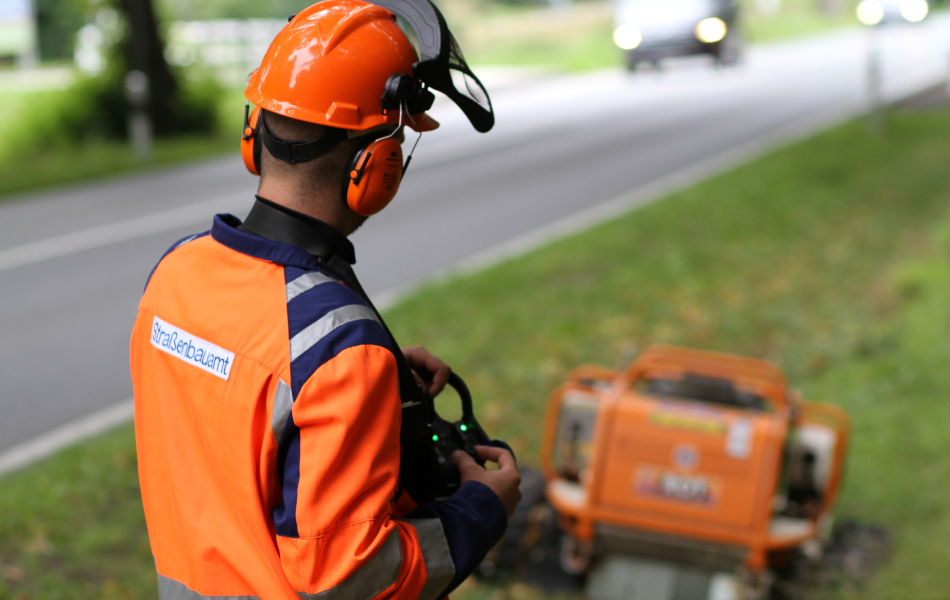 Ein Mann in orangener Arbeitskleidung steht am Straßenrand und steuert mit einer Fernbedienung einen Mähroboter, der das Grün an der Straße mäht. Auf der Straße fahren Autos.