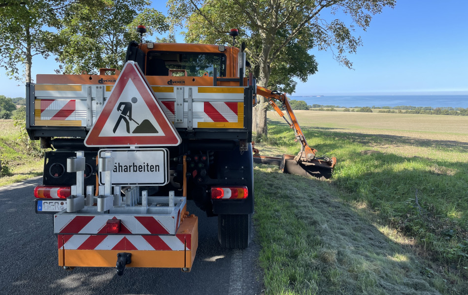 Ein orangenes Mähfahrzeug fährt am Rand einer Straße und hat seine beiden Mäharme in unterschiedlicher Länge ausgefahren. Es mäht die Grünfläche am Straßenrand.