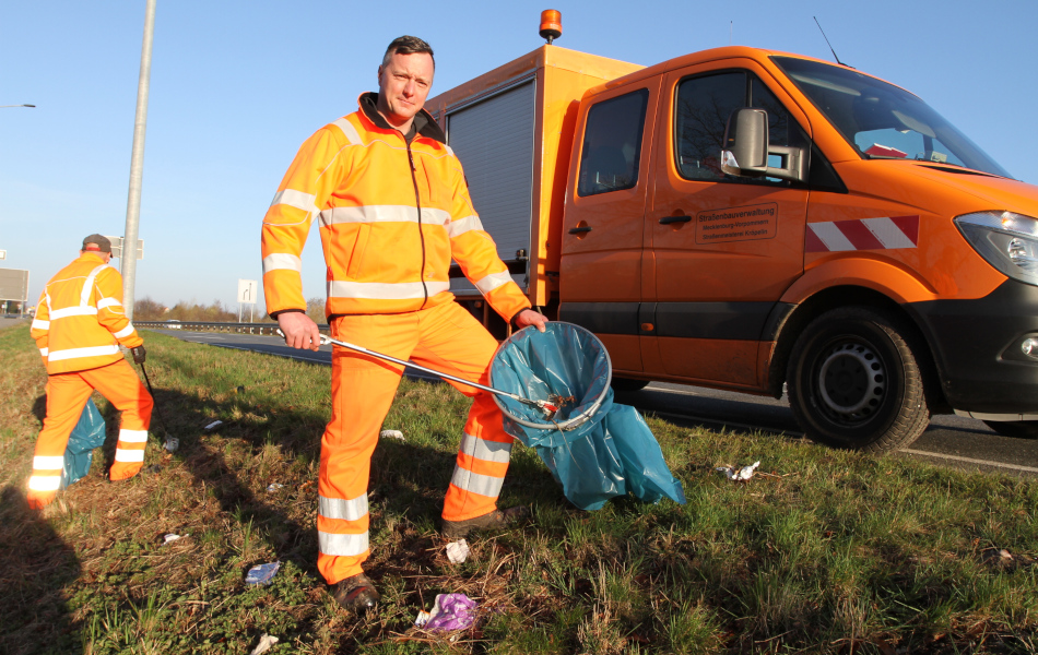 Ein Mann in orangener Arbeitskleidung hebt mit einem Greifgerät Verpackungsmüll von einer Grünfläche am Straßenrand auf und legt den Müll in einen blauen Sack. Im Hintergrund sammelt ein weiterer Arbeiter Müll und auf der Straße steht ein orangenes Arbeitsfahrzeug.. 