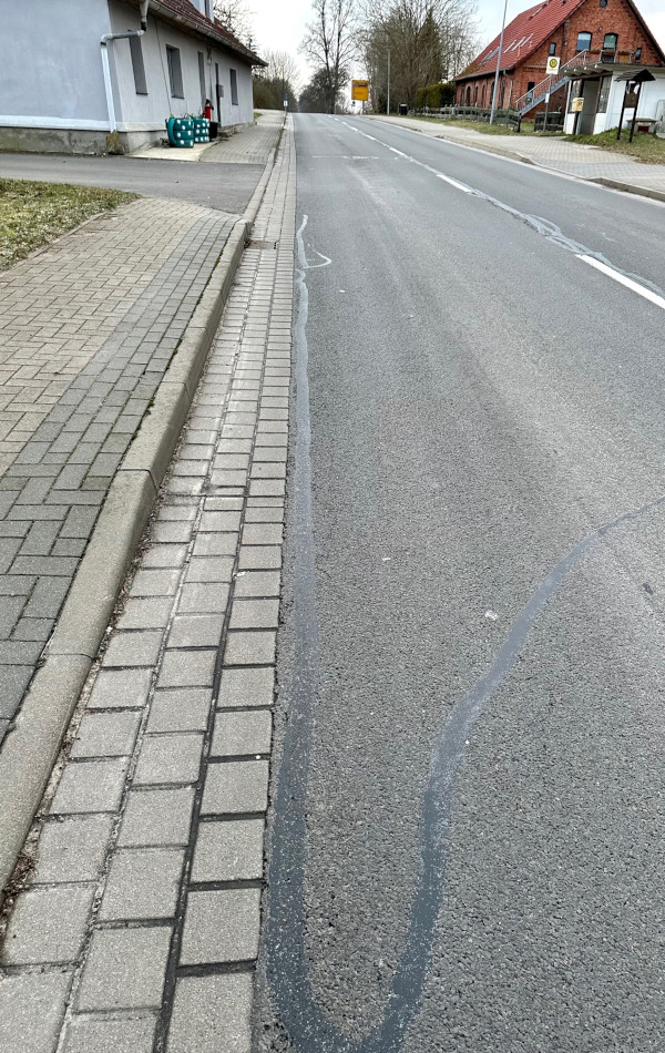 Eine Straße führt durch eine Ortschaft. Links an der Straße führt ein gepflasterter Gehweg entlang. Auf dem Asphalt der Straße befindet sich ein langer, schmaler, gummiartiger Streifen.