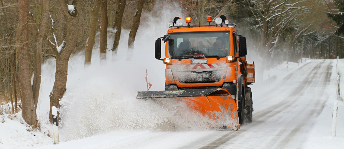 Ein orangenes Fahrzeug ist auf einer schneebedeckten Straße in einem Wald unterwegs und schiebt mit einem Pflug den Schnee zur Seite.