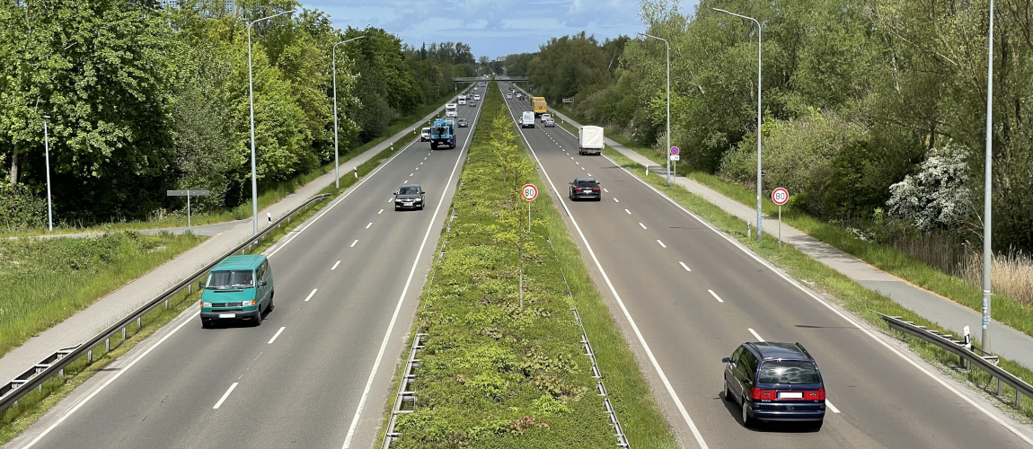 Autos fahren auf einer zweibahnigen Straße. Die beiden Fahrbahnen sind durch einen Grünstreifen in der Mitte getrennt.