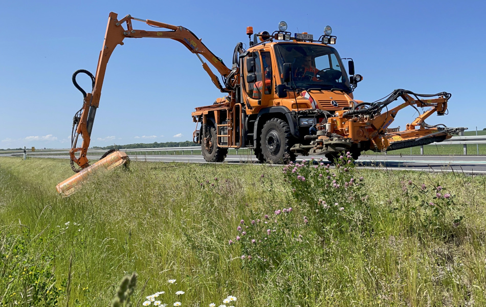 Ein orangenes Mähfahrzeug fährt auf einer Straße und mäht mit seinem ausgefahrenen Auslegearm das Gras am Straßenrand ab. (Interner Link: Interner Link)