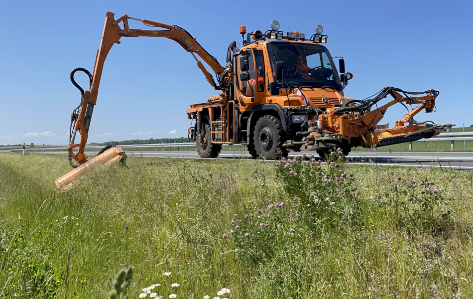 Ein orangenes Mähfahrzeug fährt auf einer Straße und mäht mit seinem ausgefahrenen Auslegearm das Gras am Straßenrand ab.