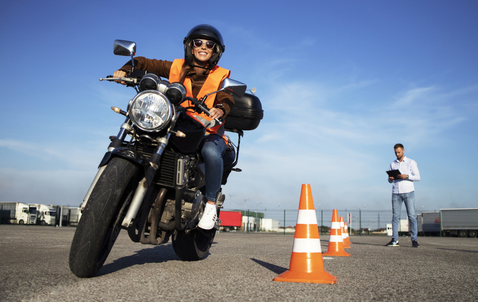 Eine Studentin mit Helm nimmt Motorradunterricht und übt das Fahren. Im Hintergrund sind Verkehrskegel und ein Fahrschullehrer mit einer Checkliste für die Bewertung und Auswertung der Fahrt. © iStock.com/Smederevac Eine Studentin mit Helm nimmt Motorradunterricht und übt das Fahren. Im Hintergrund sind Verkehrskegel und ein Fahrschullehrer mit einer Checkliste für die Bewertung und Auswertung der Fahrt.