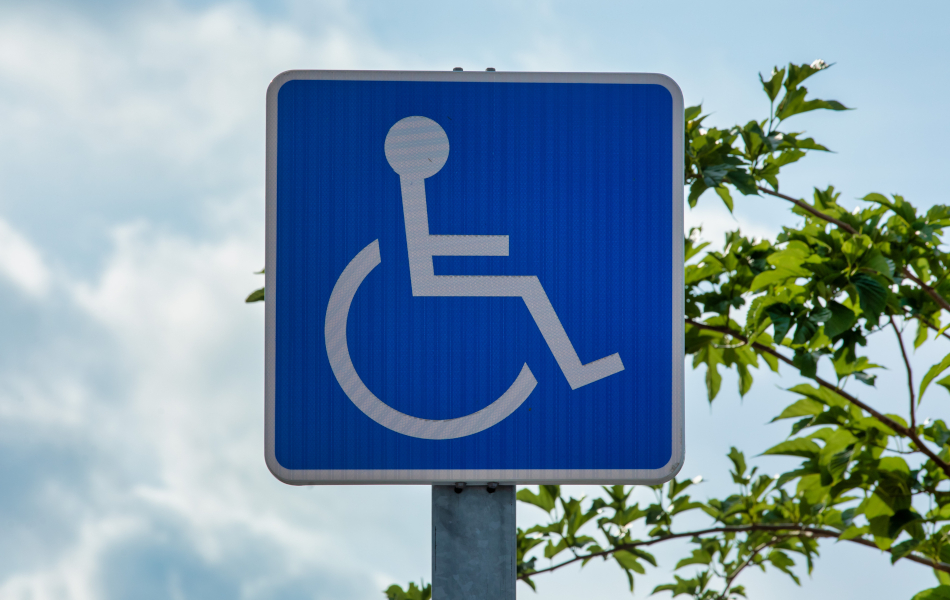Blaues Behindertenparkplatzschild mit weißem Rollstuhl-Symbol vor leicht bewölktem Himmel und einer Baumkrone © iStock.com/Peter Togel Blaues Behindertenparkplatzschild mit weißem Rollstuhl-Symbol vor leicht bewölktem Himmel und einer Baumkrone